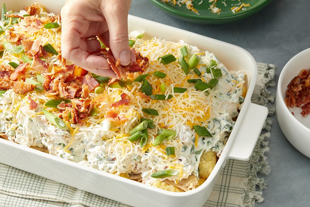 A hand sprinkles crispy bacon bits on top of a loaded potato casserole in a white baking dish. The dish is topped with shredded cheese, chopped green onions, and crispy bacon on a textured surface with a green napkin.
