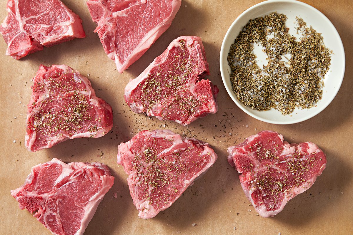 Overhead view of seasoning the lamb loin chops with the spice rub on brown parchment paper, with the spice rub kept in a bowl next to it.