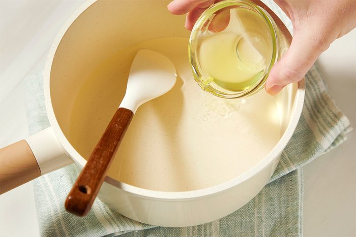 A hand pouring limoncello from a small glass bowl into a saucepan containing a sugar and water with a spoon, placed on a cloth.