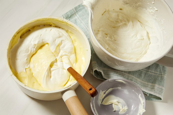 Bowls of fluffy whipped cream on a textured cloth, with a wooden-handled spatula mixing in one of the bowls.