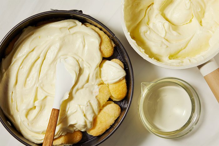 A baking tin is filled with ladyfingers and creamy mixture, being spread with a spatula. Nearby is a bowl of the same cream and a small jug of limoncello.