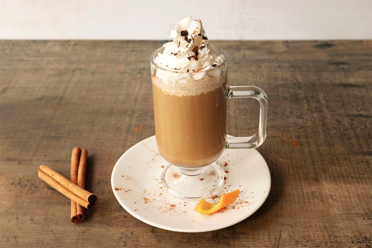 A glass mug of coffee topped with whipped cream and chocolate shavings sits on a white plate with a wooden table. Two cinnamon sticks and a small orange peel are placed on the plate beside the mug.