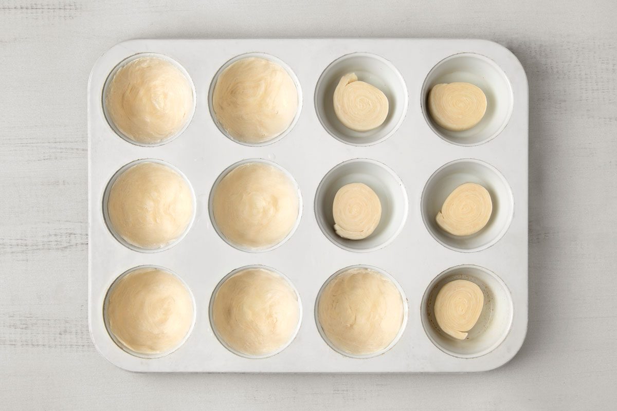 overhead shot of a white muffin tin with twelve empty muffin cups; the muffin tin sits on a white wooden surface, the cups are filled with unbaked pie crusts; some of the cups have flat disks of pie crust in them and some of the cups have a hollowed out pie crust ready to be filled with filling