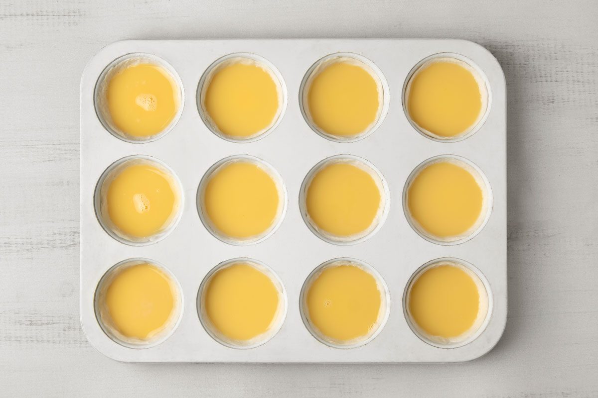 overhead shot of a white, rectangular baking tray with twelve circular depressions filled with a yellow custard-like mixture; the tray is sitting on a white countertop with subtle grey streaks