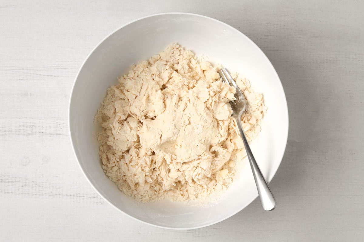 overhead shot of a white bowl with a mixture of dough and flour in it, a silver fork is lying on top of the dough, the bowl is on a white wooden table;