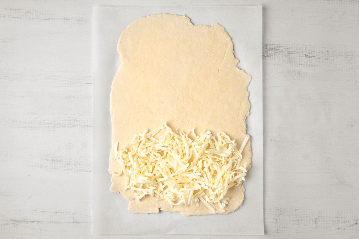 overhead shot of a sheet of parchment paper lies on a white wooden surface; on top of the parchment paper is a disc of dough that is covered in shredded butter
