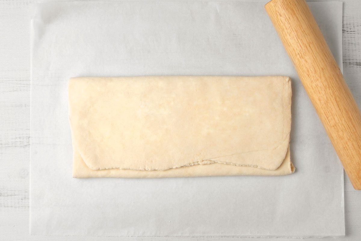 overhead shot of a piece of rolled-out dough on a piece of parchment paper, a wooden rolling pin lies to the right of the parchment paper; The dough is a pale beige color, and it is rectangular in shape