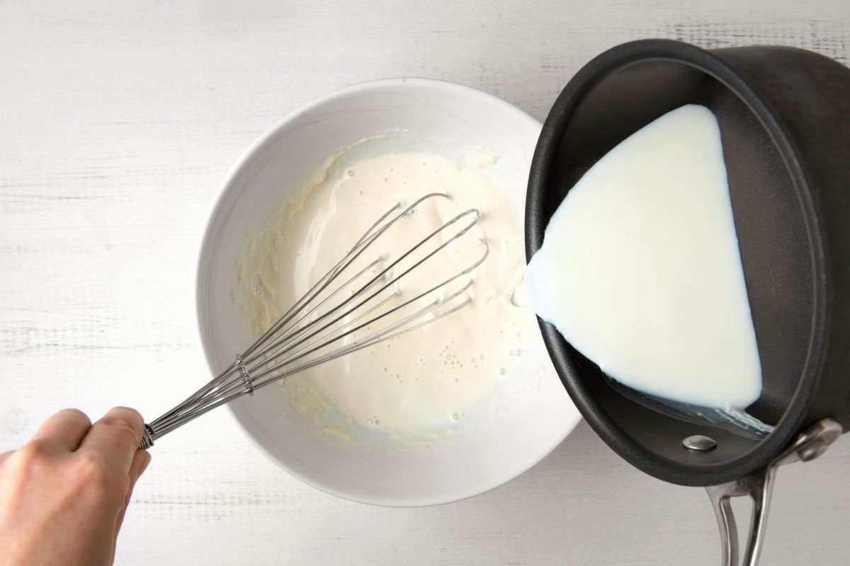 overhead shot of a person is pouring milk from a black skillet into a white bowl, a whisk is in the bowl, the milk is already partially in the bowl, the person is holding the handle of the whisk, the whisk is close to the edge of the bowl