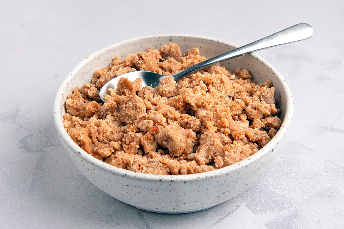 A speckled white bowl filled with crumbly brown sugar sits on a light gray marble surface. A silver spoon is resting in the sugar, angled slightly to the right.