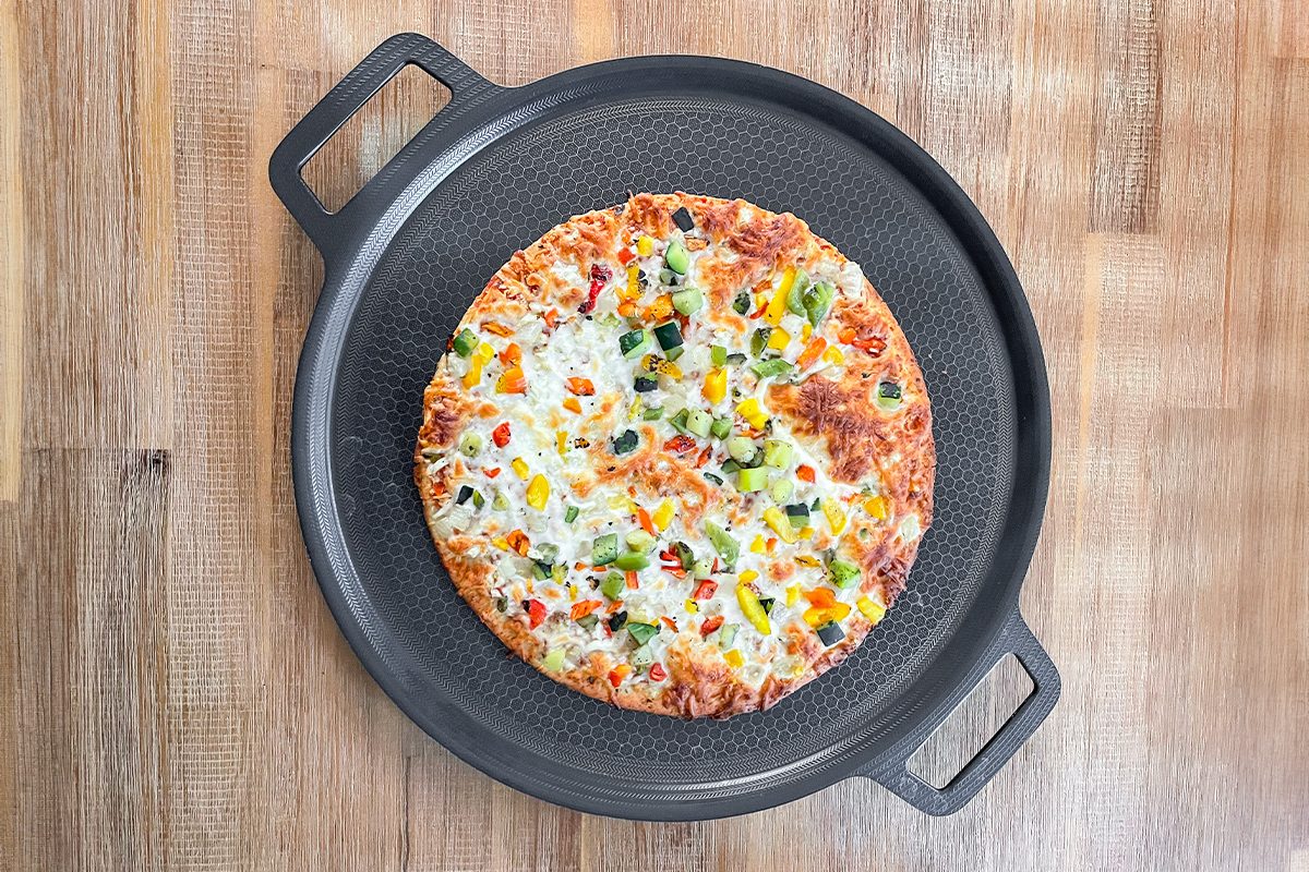 A baked pizza topped with colorful vegetables, including green peppers, red onions, and sweetcorn, sits on a black perforated pizza pan placed on a wooden surface.