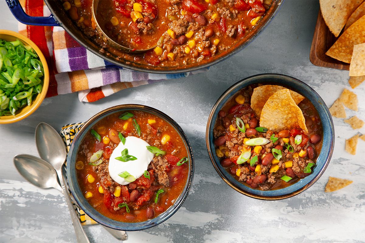 Two bowls of hearty chili topped with sour cream, green onions, and tortilla chips. A pot of chili and more chips are nearby, with spoons and a colorful napkin. The chili contains meat, corn, beans, and tomatoes.