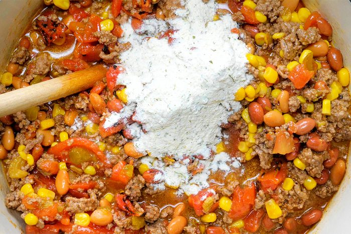 A pot of colorful chili is being prepared, featuring ground meat, kidney beans, corn, diced tomatoes, and green peppers. A heap of white flour is piled in the center, with a wooden spoon resting on the side.