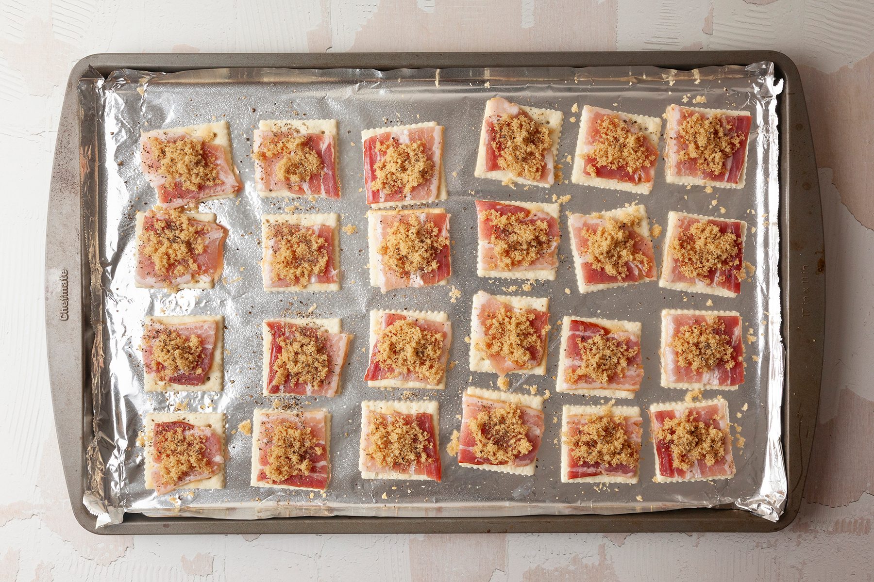 Baking sheet lined with foil, featuring 24 saltine cracker-based appetizers. Each cracker is topped with a slice of meat and sprinkled with a breadcrumb mix, ready to be baked.