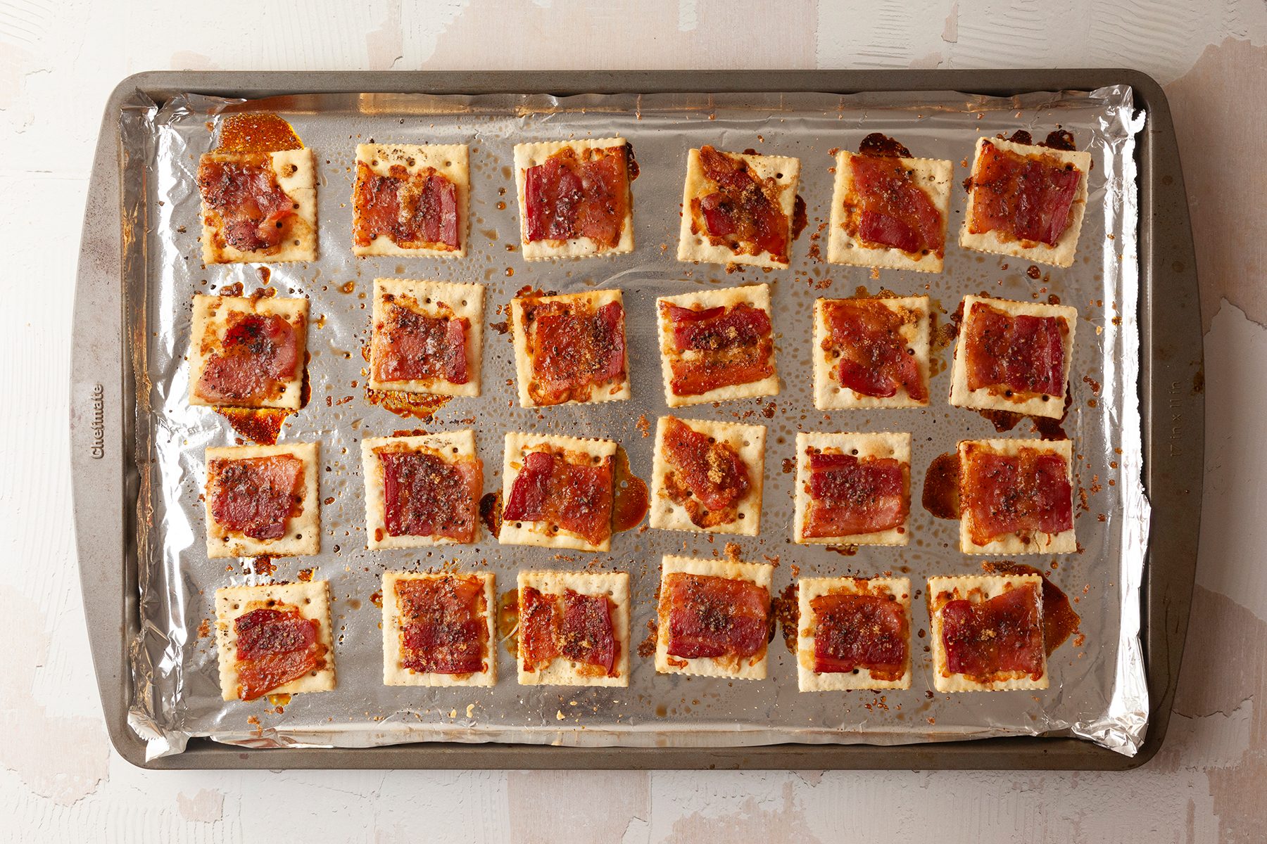 A baking tray covered with foil holds evenly spaced crackers topped with bacon and a caramelized glaze. The crackers are arranged in a 5x5 grid and are baked until golden brown.