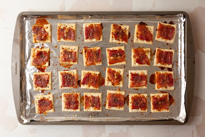 A baking tray covered with foil holds evenly spaced crackers topped with bacon and a caramelized glaze. The crackers are arranged in a 5x5 grid and are baked until golden brown.