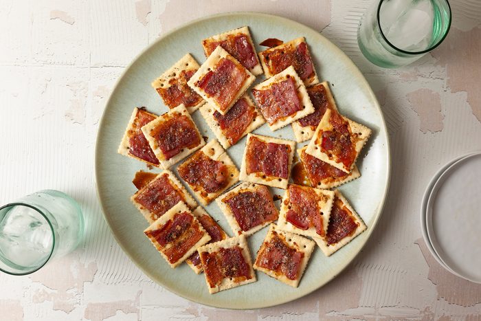 A green plate filled with crispy crackers topped with a spread resembling bacon jam. The plate is set on a textured, light-colored surface. Two glasses with ice and a stack of white plates are partially visible around the plate.