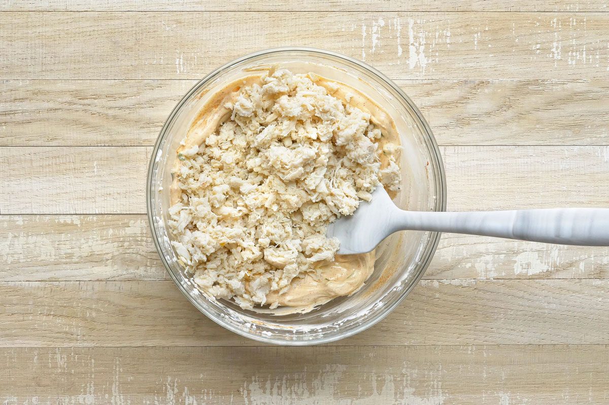 overhead shot of a glass bowl containing a mixture of creamy sauce and flaked crabmeat; a white spatula is sticking out of the bowl; the bowl is sitting on top of a wooden table