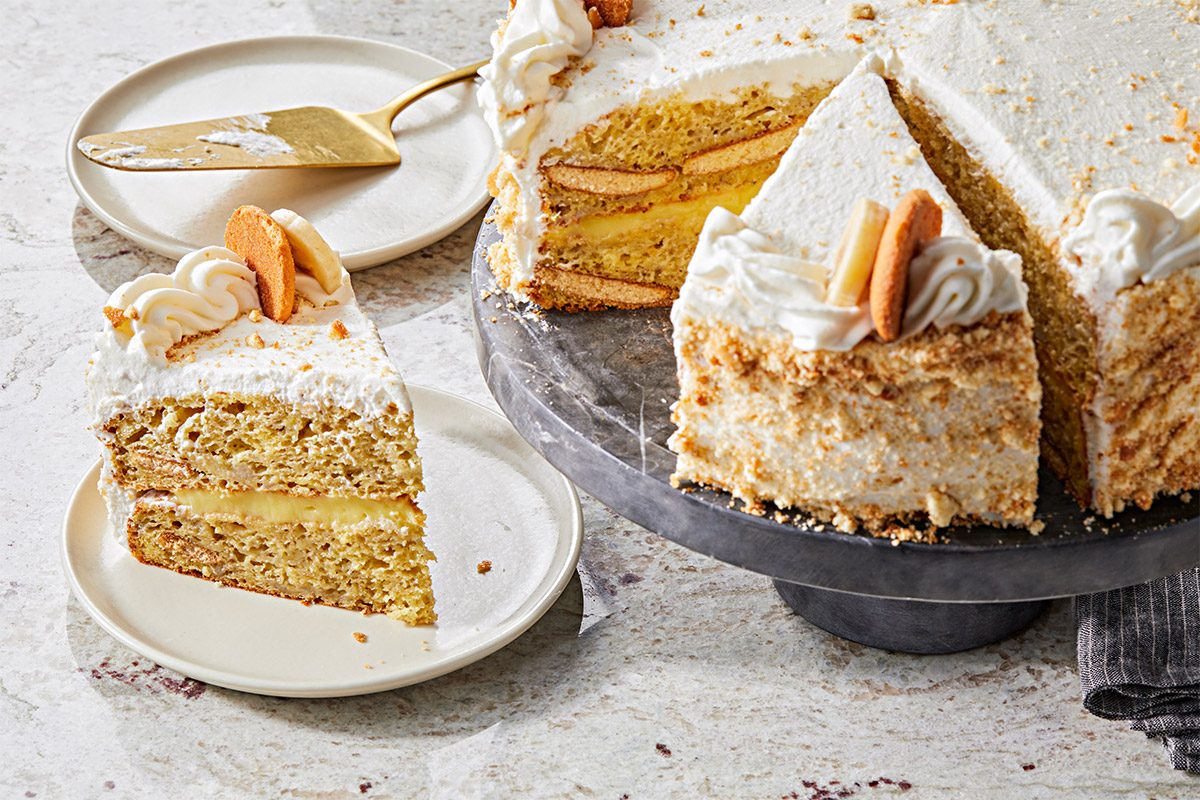 3/4th shot of a banana pudding cake that is presented on a round cake stand; next to the cake, a slice is displayed on a plain white plate, the cake is set against a speckled light colored surface