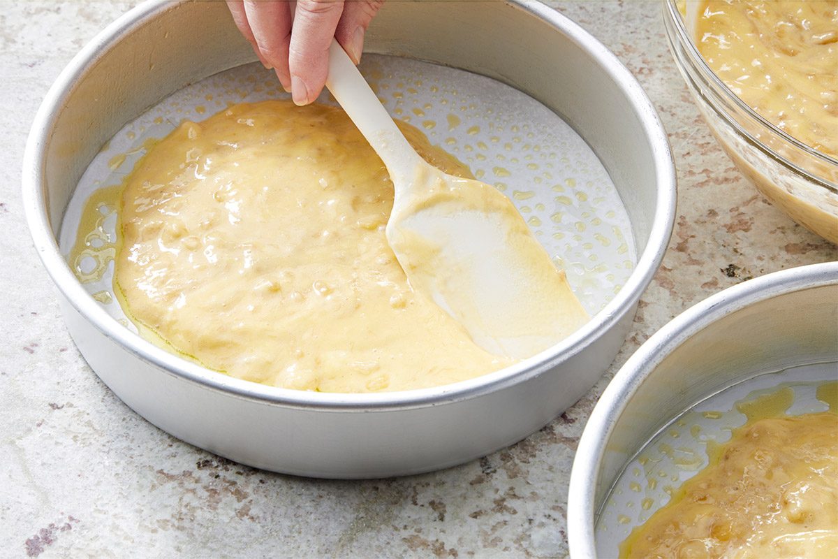 3/4th shot of a hand using a spatula to spread a creamy batter into a round cake pan, which is set on a textured surface, Nearby, there is another round cake pan and a bowl containing more of the batter