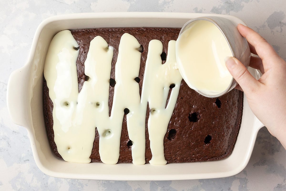 A hand pouring creamy white sauce over a chocolate cake with holes in a rectangular baking dish. The dish is on a light marble surface. The sauce is being added to half of the cake, seeping into the holes.