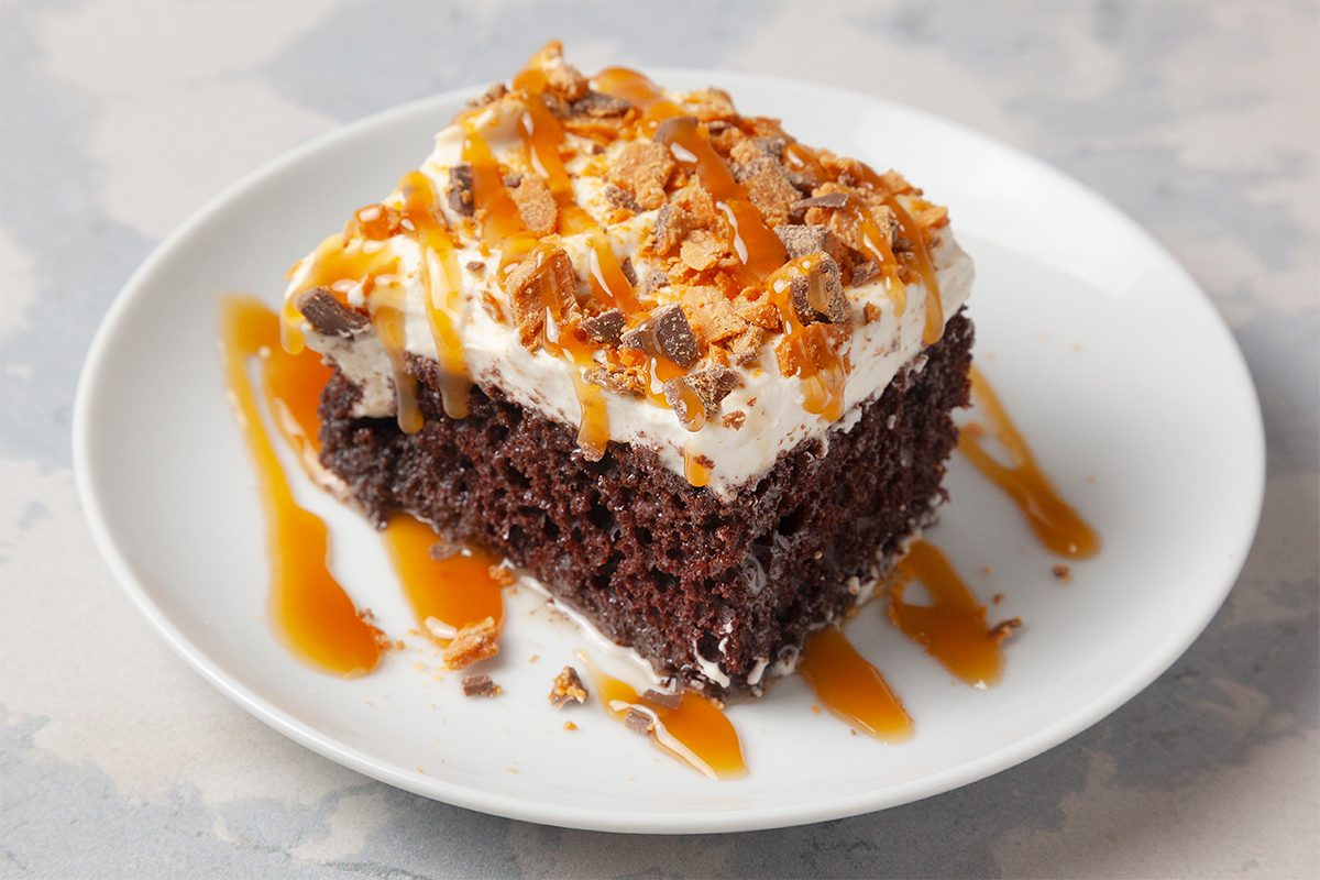 A slice of chocolate poke cake on a white plate, topped with whipped cream and sprinkled with crushed toffee. Caramel sauce is drizzled over the top and the plate. The background is a light gray surface.
