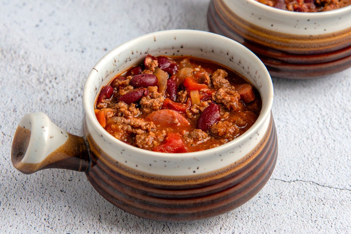 a close-up of a bowl of chili; the bowl is brown with a white rim and a handle; the bowl is sitting on a light gray countertop