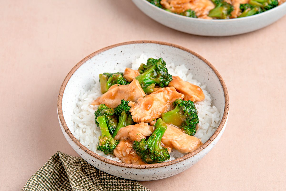 A bowl of white rice topped with stir-fried chicken and broccoli in a light sauce. The dish is served in a ceramic bowl on a beige surface, with part of another bowl visible in the background. A folded cloth napkin is placed nearby.