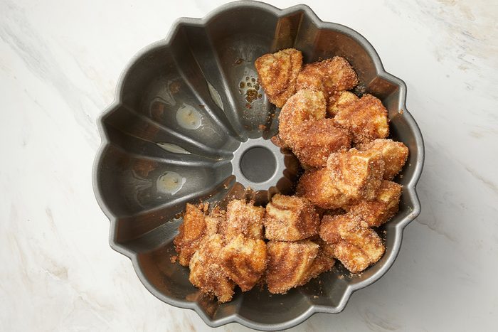 overhead shot of a partially filled bundt pan on a light colored countertop; inside, there are clusters of golden brown, doughy pieces coated in a sugary, cinnamon mixture; the countertop has a subtle marble pattern