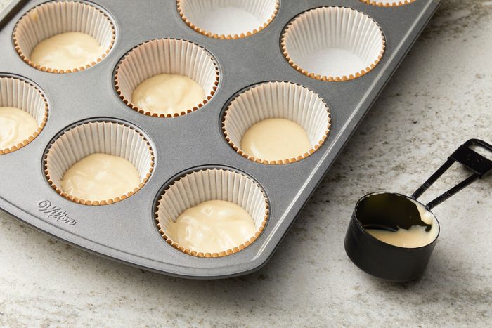a closeup, top down view of a muffin tin filled with cupcake liners and batter; ten of the depressions are lined with white paper liners, and eight of those liners are filled with a light colored batter; in the lower right corner, a black plastic measuring cup containing some of the same light colored batter is visible; the countertop is a light gray with a subtle speckled pattern
