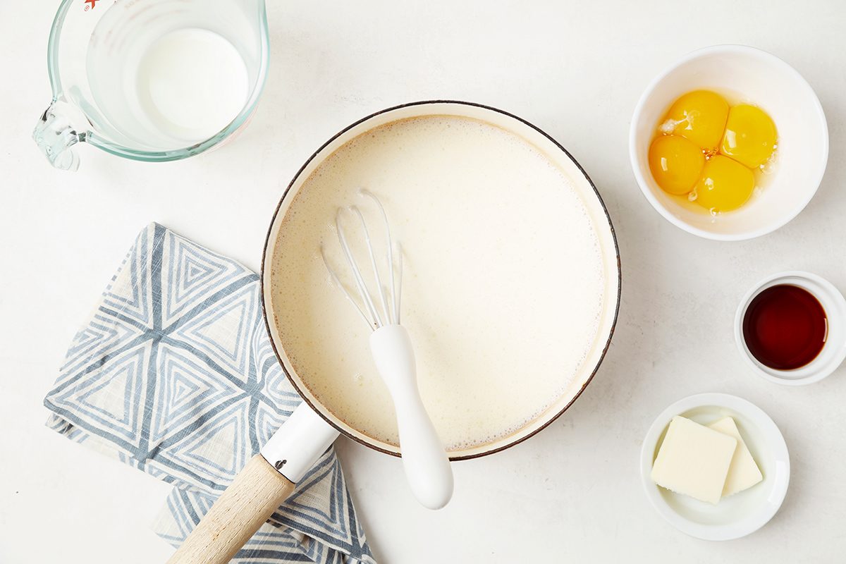 A mixing bowl with creamy batter and a whisk is surrounded by a measuring cup of milk, a bowl with three egg yolks, a small bowl of vanilla extract, a dish of butter, and a blue patterned cloth on a white surface.