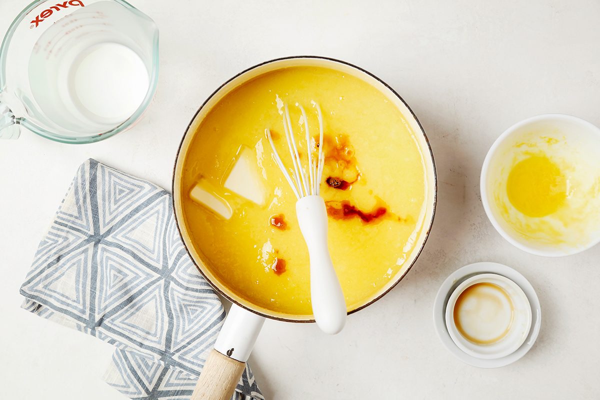 A pot of yellow custard with a whisk inside, on a light countertop. Next to it are a measuring jug, a small bowl with melted butter, and an empty bowl on a patterned cloth.