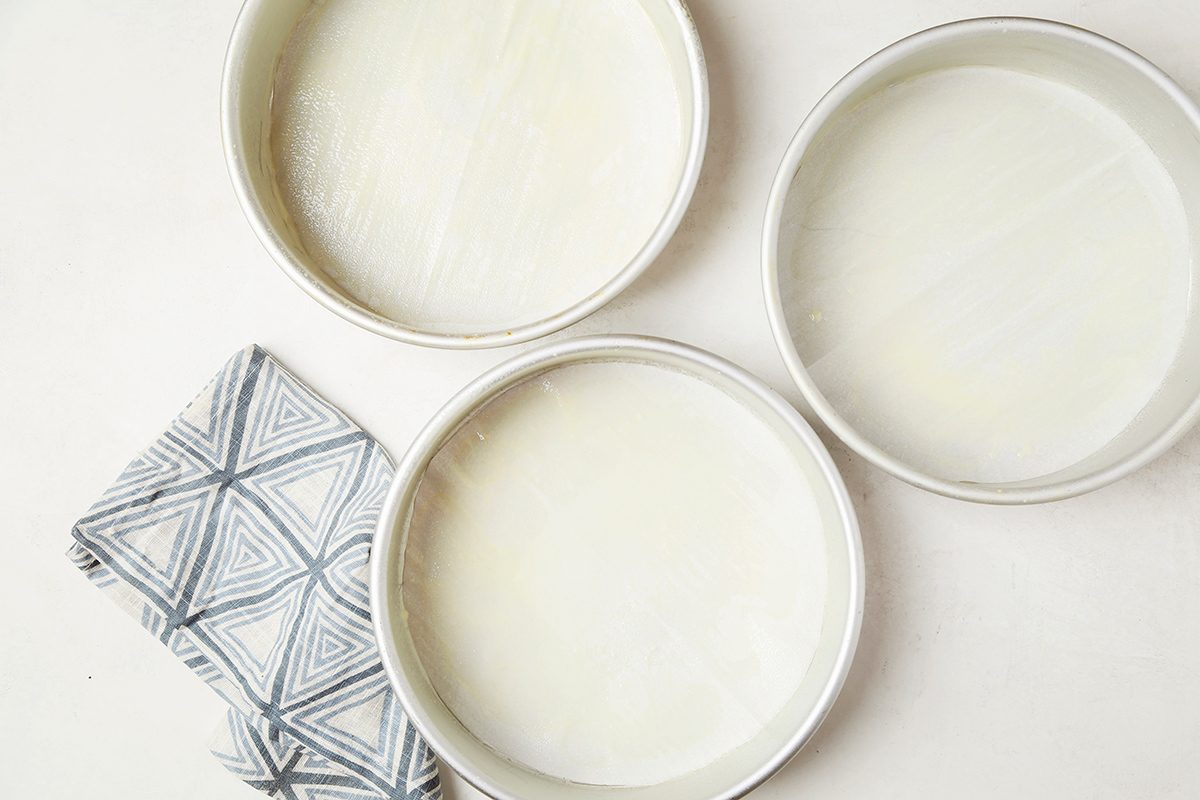 Three empty round baking pans with parchment paper lining at the bottom are arranged on a light surface. A folded cloth with a geometric pattern lies beside the pans.