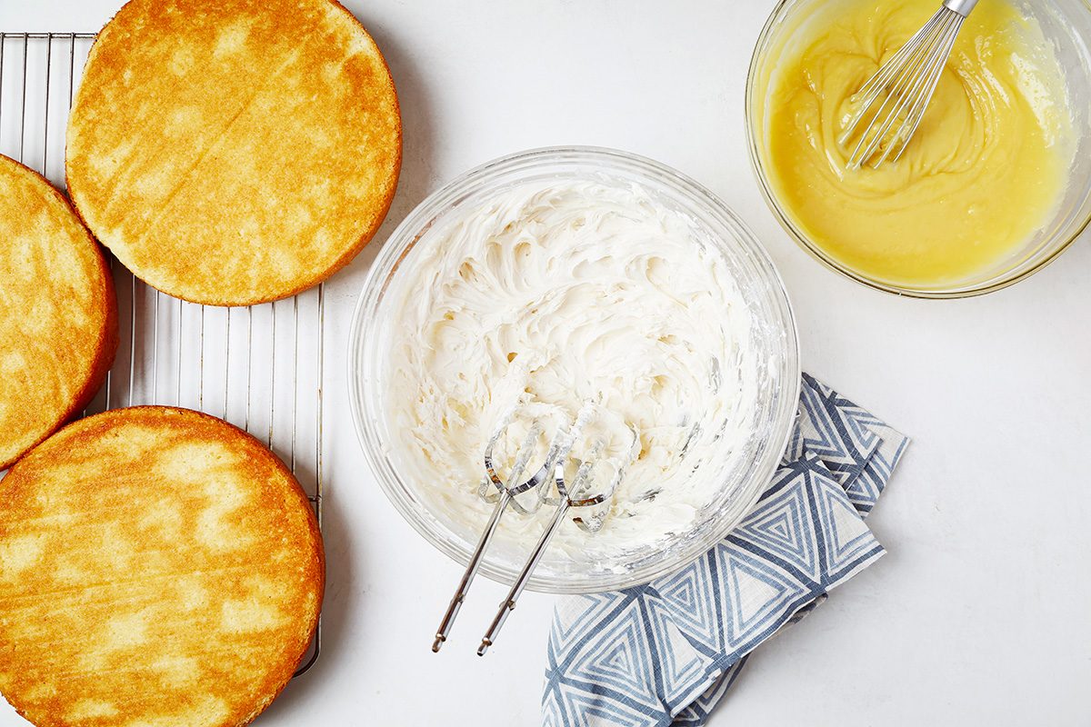 Three round cake layers cool on a wire rack. Nearby, a bowl of whipped cream with a mixer and a bowl of yellow batter with a whisk sit on a patterned cloth. The scene is set on a white surface.