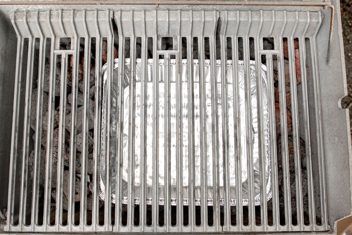 overhead shot of a barbecue grill with a silver aluminum pan resting on the grill grates; the pan is surrounded by charcoal briquettes and the grates are made of metal