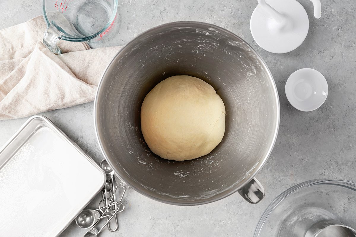 A ball of dough rests in a stainless steel mixing bowl on a gray countertop. Nearby are a measuring cup with water, a white cloth, measuring spoons, a baking tray, and an empty white cup.
