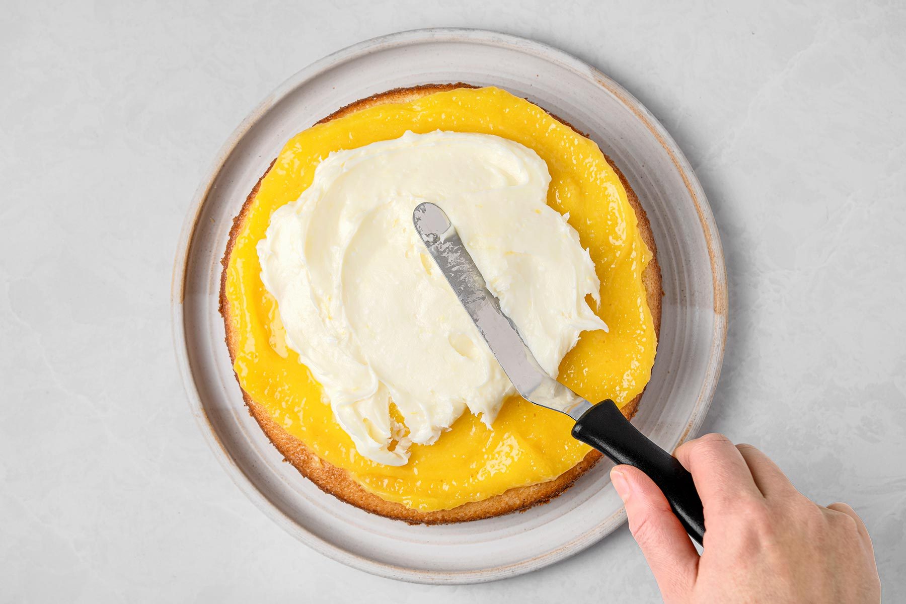 Spread cream mixture over cake kept on top of a grey marbletop.