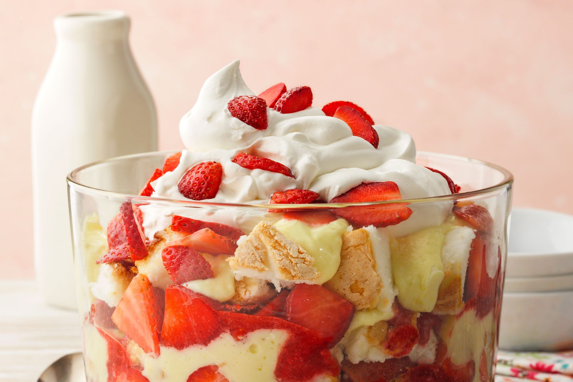 Strawberry Lemon Trifle; low camera angle; white washed wood surface; light wood surface; russian surface; trifle bowl; strawberries; lemon pudding; angel food cake; whipped topping; cool whip; lemon zest; milk bottle; white bowl; spoon