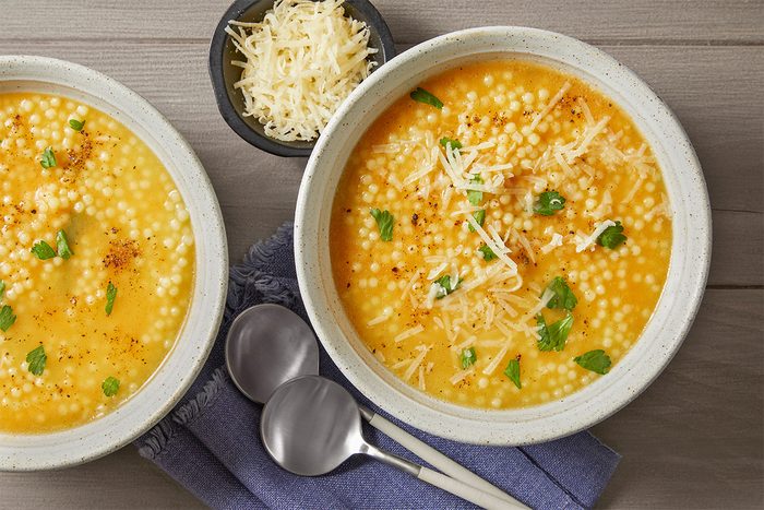 Two bowls of creamy soup topped with small pasta, herbs, and grated cheese are on a wooden table. Beside them are two spoons on a blue napkin and a small bowl of extra grated cheese.