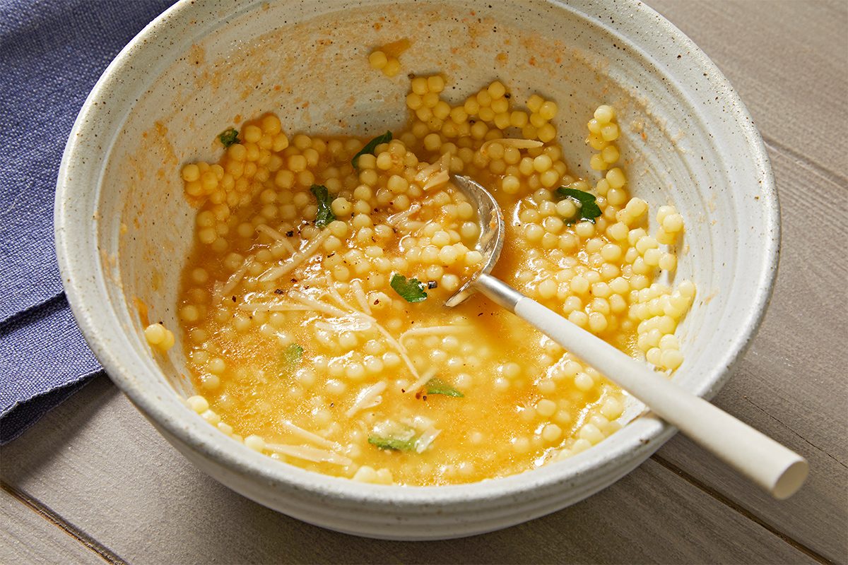A bowl of couscous soup with small pasta, garnished with herbs, is set on a wooden surface. A spoon rests in the bowl, and a folded blue napkin is nearby. The soup has an orange hue.