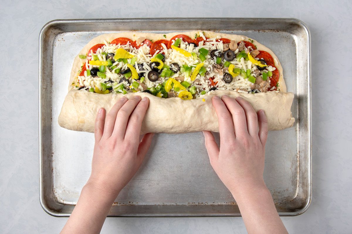 overhead shot of a pair of hands rolling up a rectangular pizza on a baking tray, the baking tray is metallic and sits on a plain grey background;