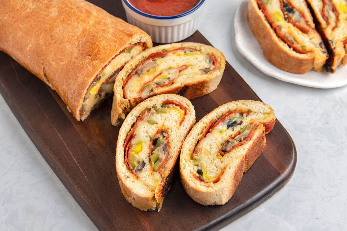 3/4th shot of a Pepperoni Pizza Loaf cut into slices and arranged on a wooden board, a small bowl of red dipping sauce is placed at the top left, and a plate with additional slices is in the top right corner, the wooden board and plates are set on a light grey surface;