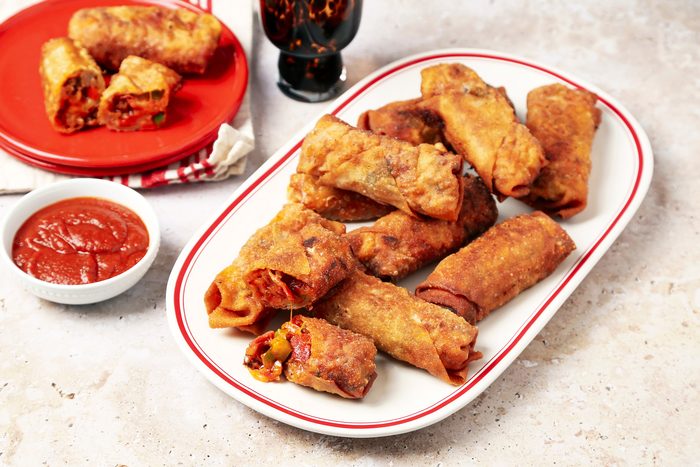Taste of Home's Pizza Egg Rolls shot on a beige marble surface. Pizza filling inside egg roll wrappers and fried until golden brown and crispy and served with pizza sauce for dipping.