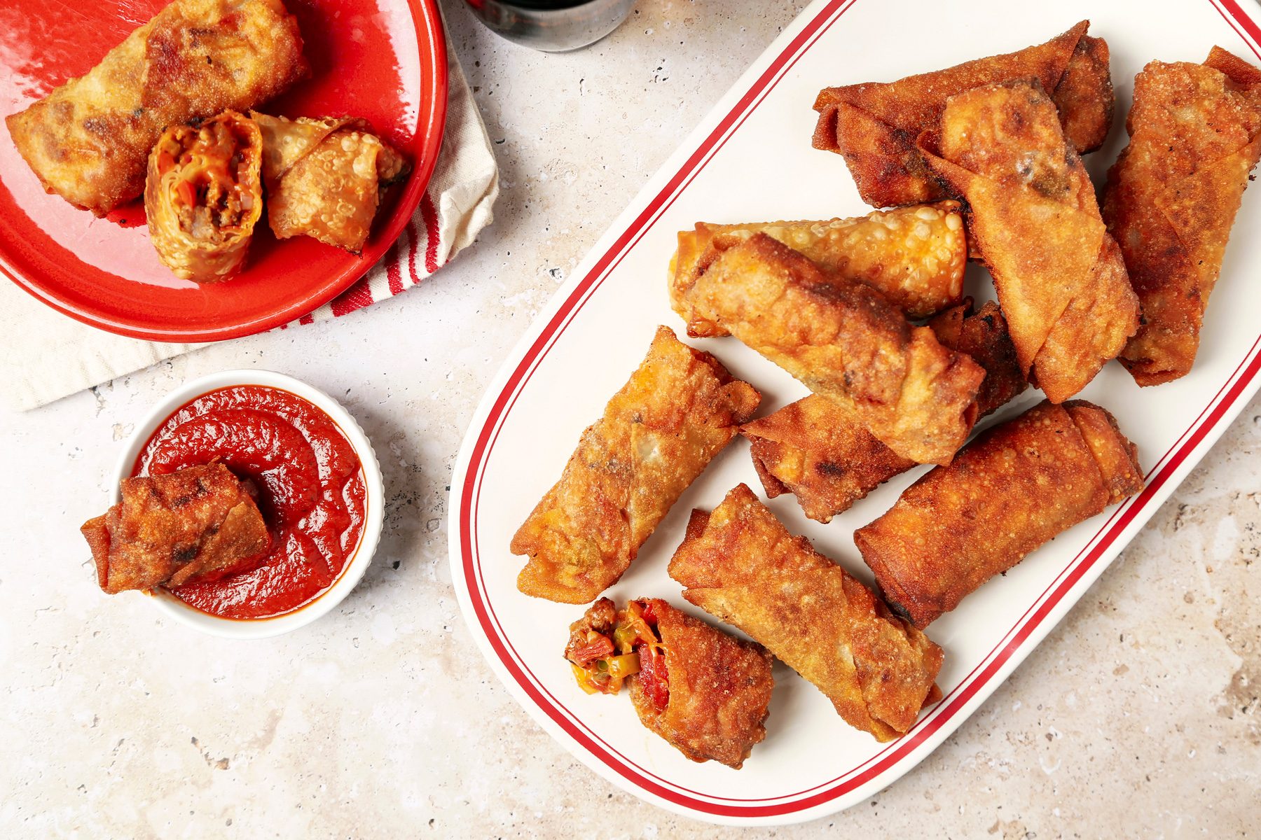 Taste of Home's Pizza Egg Rolls shot on a beige marble surface. Pizza filling inside egg roll wrappers and fried until golden brown and crispy and served with pizza sauce for dipping.