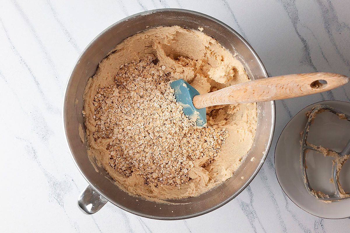 A metal mixing bowl on a marble countertop contains cookie dough with oats being mixed using a blue spatula.