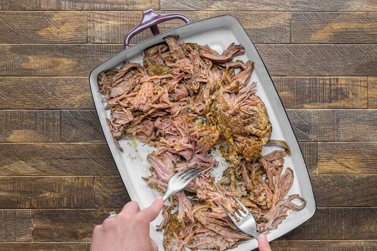 overhead shot of a white baking dish with a purple handle filled with shredded meat; the dish is sitting on a dark brown wooden surface; a person is using two forks to pull apart the shredded meat