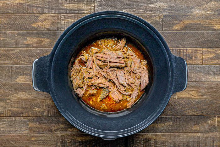 overhead shot of a black slow cooker filled with shredded meat in a brown sauce, it is set on a wooden surface