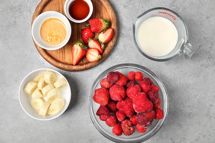 A wooden plate with honey, vanilla, and halved strawberries; a bowl with sliced bananas; a glass container with milk; and a bowl of frozen mixed berries are arranged on a gray countertop.