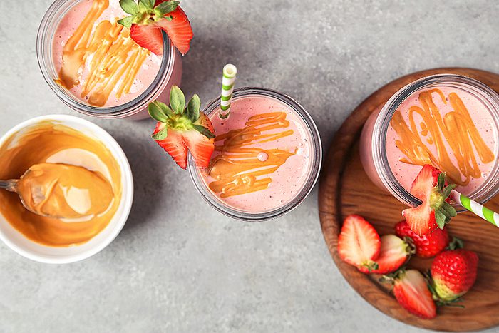 Two glasses of strawberry smoothies with striped straws and drizzled peanut butter on top. A small bowl of peanut butter with a spoon is on the side, accompanied by a wooden plate with fresh strawberries. All on a gray surface.