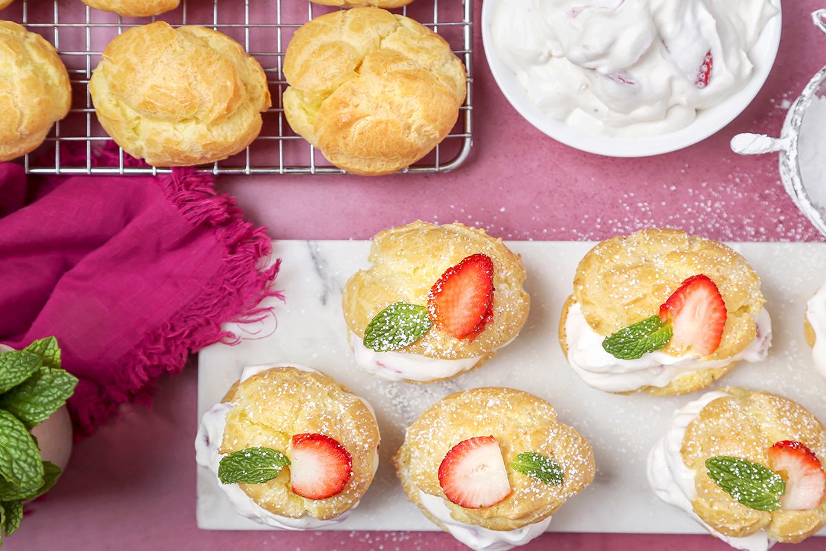 Overhead view of garnished strawberry cream puffs served next to the puffs kept on a cooling tray, cream, and a pink napkin on a pink tabletop.