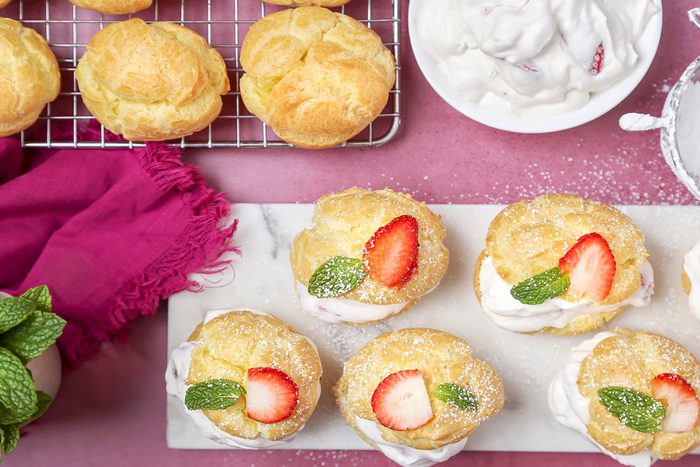 Overhead view of garnished strawberry cream puffs served next to the puffs kept on a cooling tray, cream, and a pink napkin on a pink tabletop.